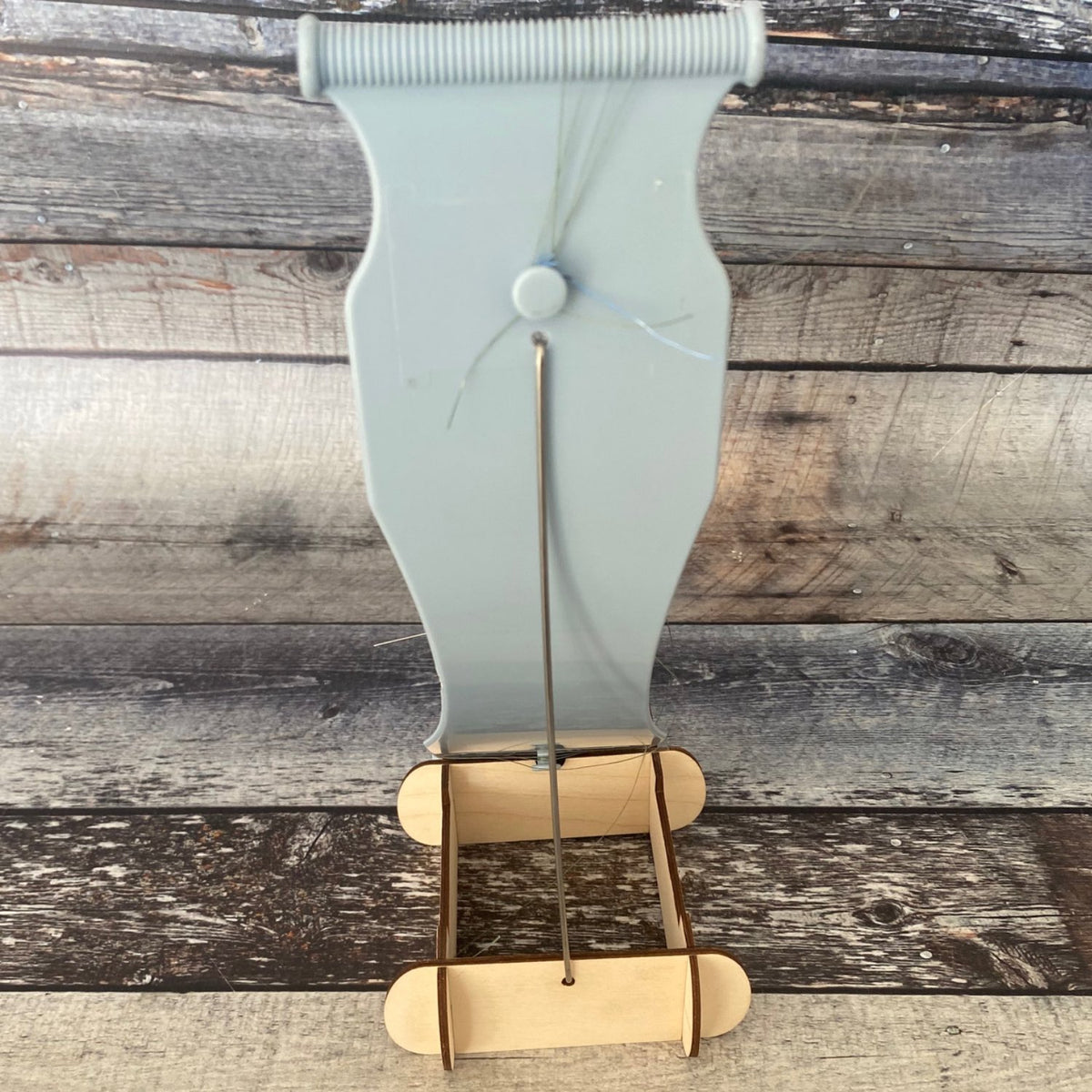 Jewel Loom Stand with Jewel Loom sitting in stand facing from the back of the loom. Metal Rod has been inserted into the back of the loom and into the back bottom hole of the Jewel Loom Stand. Photographed on faux wood background in hues of blue grey.