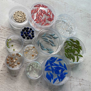 "A curated collection of jewelry making supplies from the Jewel Loom January Jewel Box Mosaic Muse kit. Features clear jars containing cobalt blue and slate geometric beads, green travertine beads, sunstone and prehnite gemstones, blue black tigers eye, and a decorative wooden button on a white wood background."