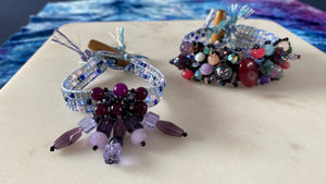 Two handmade beaded bracelets displayed on a light beige surface. The front bracelet is light blue and lavender with dangling purple beads, and the back bracelet is multicolored with red, purple, pink, and blue beads.