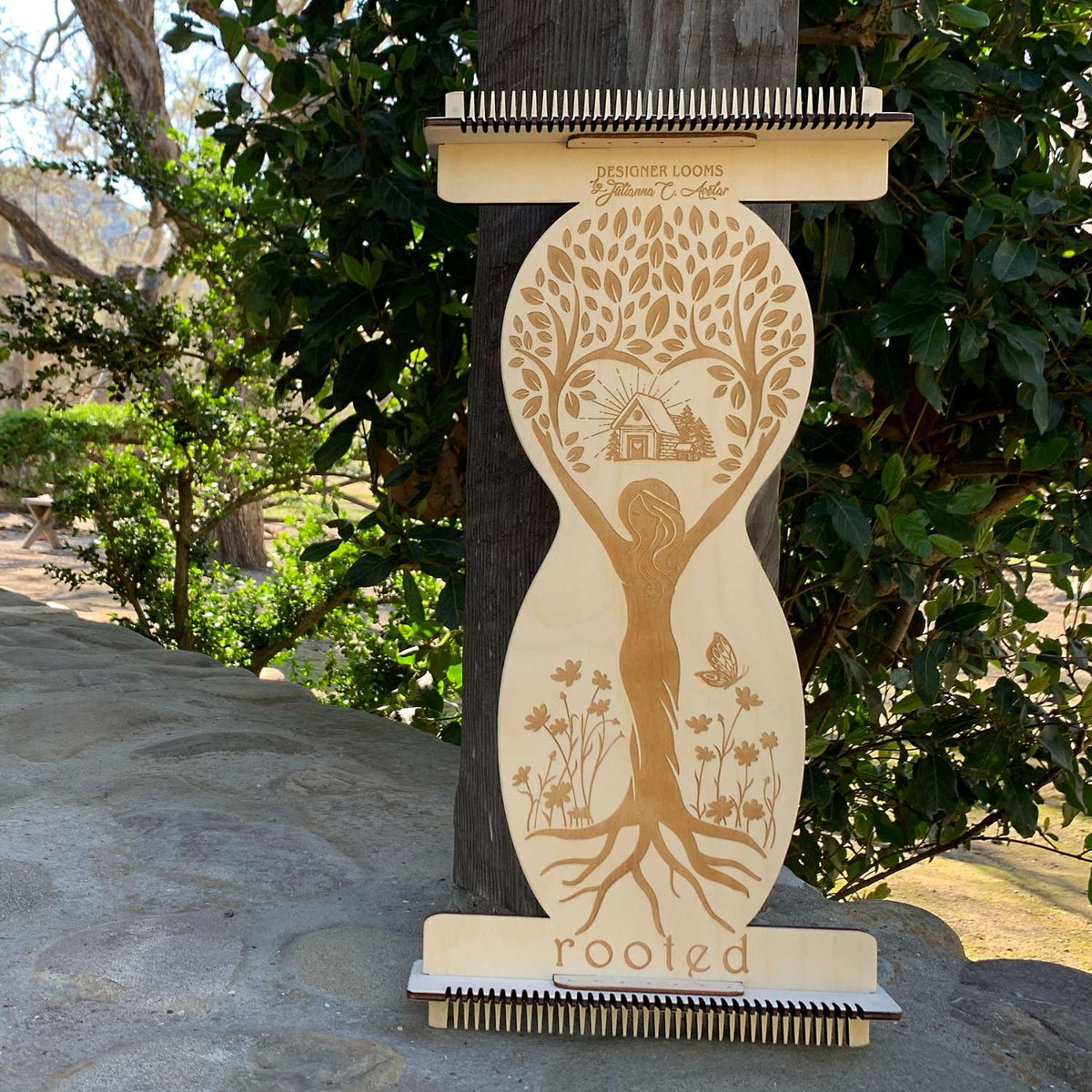 This is the ROOTED Loom made out of Sustainable Baltic Birch Wood. It is leaning up against a Wood Pillar on the front porch with a tree in the background. The ROOTED Loom is laser engraved with the silhouette of a woman holding her arms up with the image of a cottage home centered above her head. There are branches with leafs surrounding the cottage house. The branches and leafs are coming up out of the hands of the woman as if she is holding them. The bottom of the woman has roots going into the ground