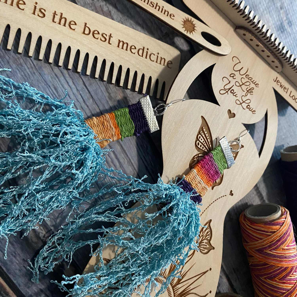 Small Sun Weaver Loom with Needle and Comb. Images of butterflies, hearts and a large sun have been laser engraved into the baltic birch wood. Wooden Needle has been engraved with the words You are my sunshine with a small sun. Wooden Comb has the words Sunshine is the best medicine with a small sun laser engraved. There is a finished pair of earrings woven from hempcord in blues, orange, yellow, pink and green along with a spool of orange varigated hempcord as a prop. The image is on a faux wood background