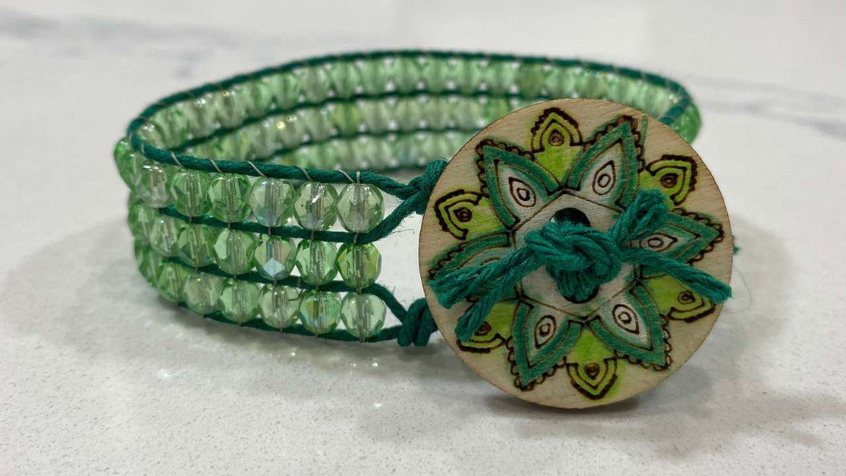 Green beaded wrap bracelet with a wooden button featuring a floral design on a white background