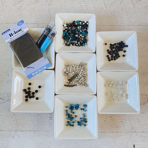 A flat-lay arrangement of craft supplies on a white wooden surface, including a package of B-Ion thread, multiple small white square dishes containing assorted beads and rhinestones in black, blue, silver, and white.
