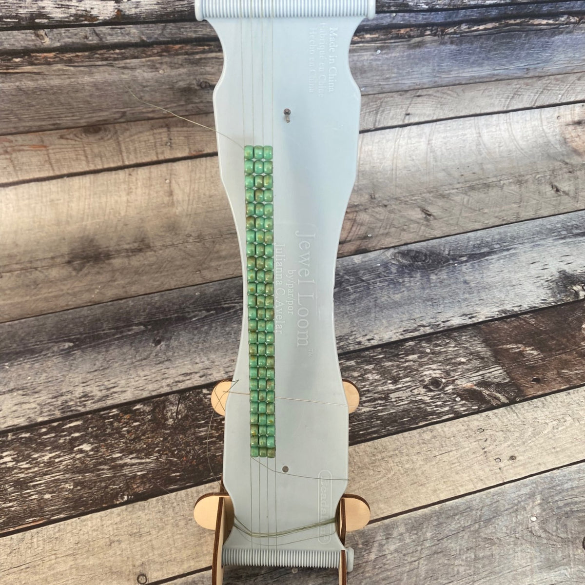 Jewel Loom Stand with Jewel Loom Bead Loom positioned in stand facing forward with example of warped loom and beads woven in as an example of how the loom sits in the stand. Image on faux wood background in blue grey tones. 