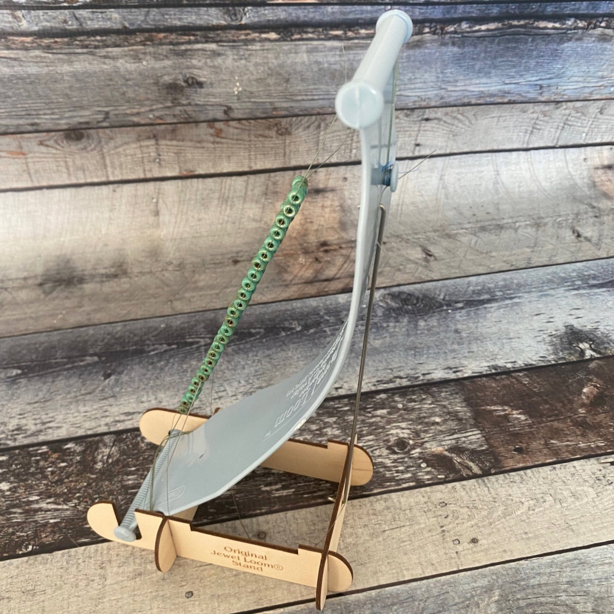 Jewel Loom Stand with Jewel Loom Bead Loom positioned in stand and facing sideways. Loom has been warped and beads have been woven in as an example of what the looms looks like from the side. Photograph has been taken on a fauz wood background in blue grey hues. 