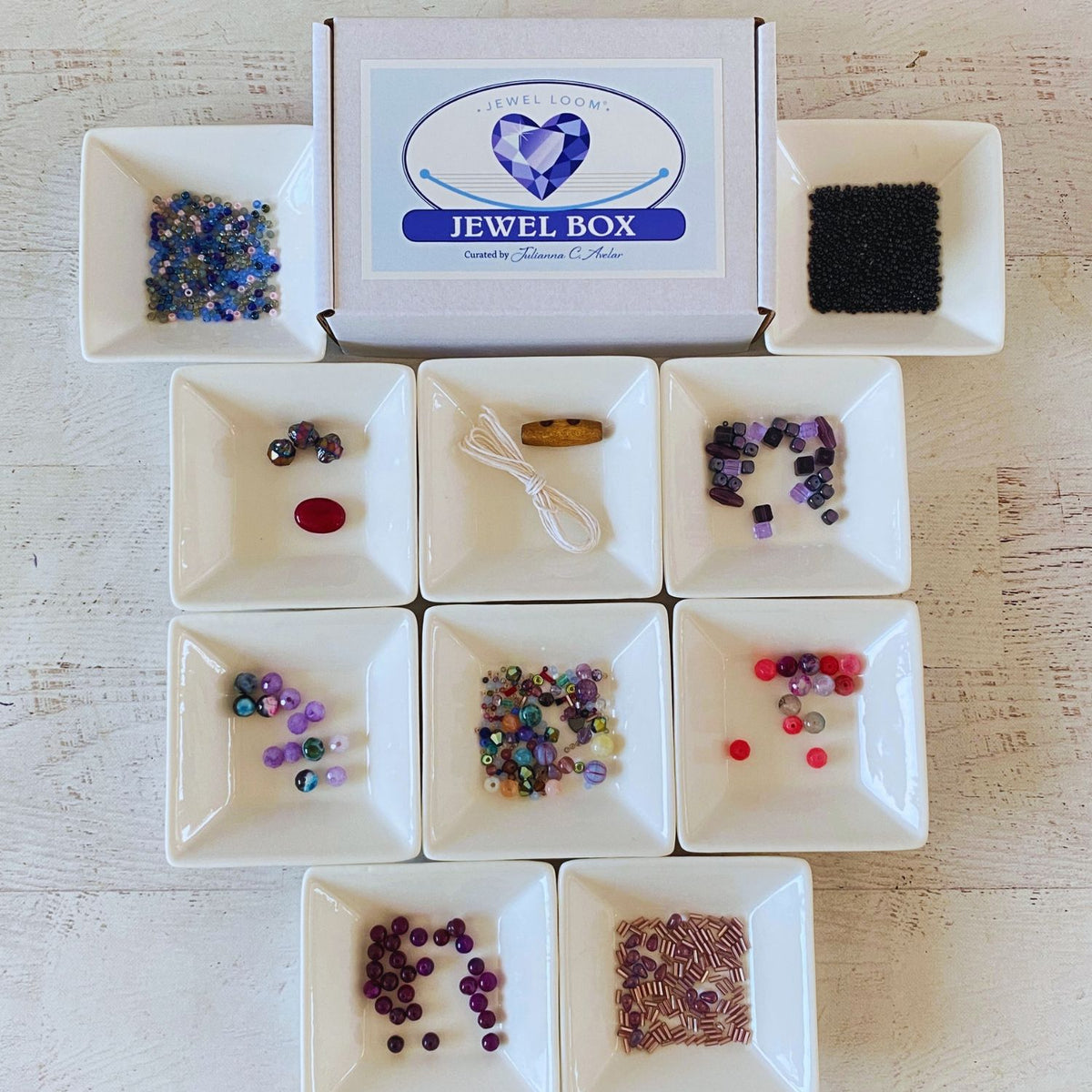 A jewelry making kit labeled 'Jewel Box' by Jewel Looms, displayed on a white wooden surface. The kit includes a white box with a blue logo, nine small white square dishes containing various colorful beads, a black mesh pad, and a wooden spool of white thread.