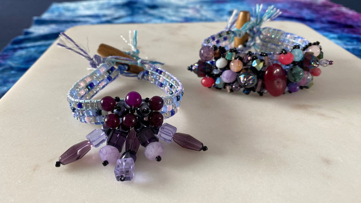 Two handmade beaded bracelets displayed on a light beige surface. The front bracelet is light blue and lavender with dangling purple beads, and the back bracelet is multicolored with red, purple, pink, and blue beads.