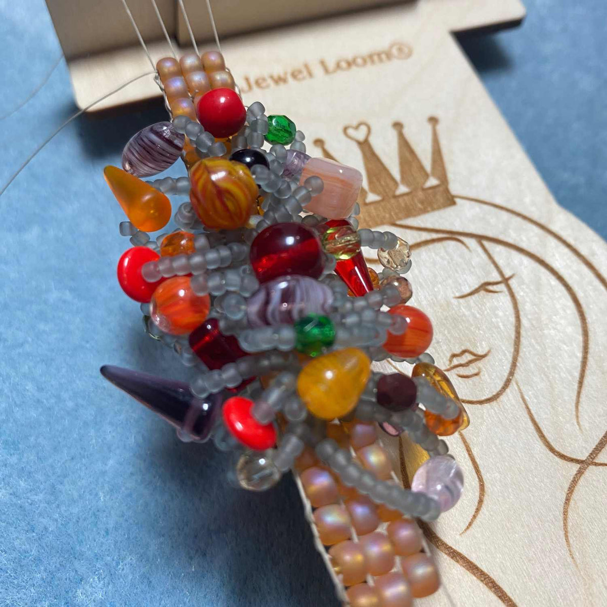 Mini Wisdom Warrior Loom with warped threads and beads woven into seed bead base in the colors of Topaz, Grey, Red, Purple, Yellow, Green and Orange. Background is in a light powder blue. 