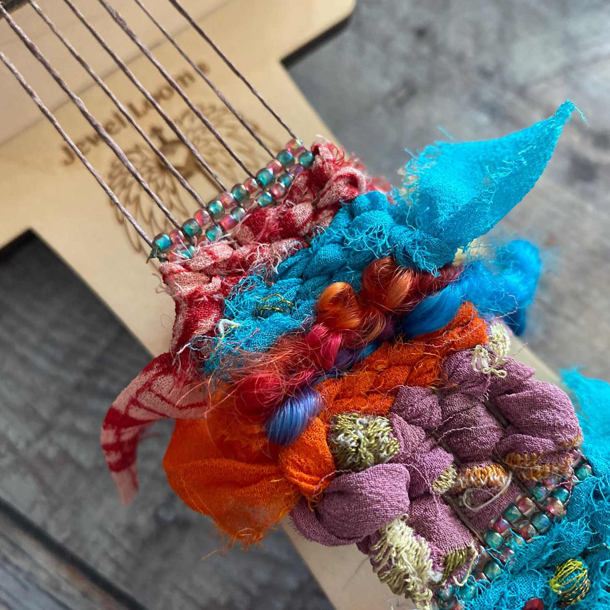 Close-up of a colorful fabric woven into the warp threads on the small wisdom warrior loom with beads on a blurred background