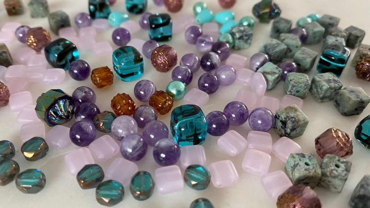A close-up, top-down view of assorted craft beads scattered across a light surface. The beads include faceted cube shapes, round cabochons, and oval beads in various colors such as purple, teal blue, pink, green, brown, and iridescent.