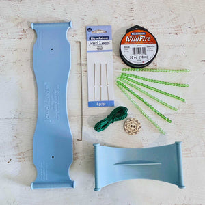Jewelry-making tools and materials including the blue Jewel Loom, Baby jewel loom, spool of wildfire thread, jewel loom needles, 6 strands of firepolish beads, hempcord and a wood button all on a wooden surface.