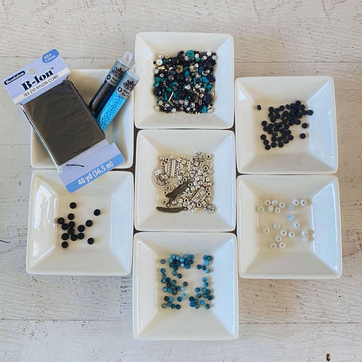 A flat-lay arrangement of craft supplies on a white wooden surface, including a package of B-Ion thread, multiple small white square dishes containing assorted beads and rhinestones in black, blue, silver, and white.
