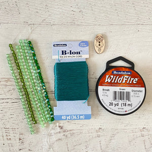 Beadalon B-Ion thread and beads laid out on a light wood surface, including green faceted beads, a spool of teal cord, a spool of green Beadalon WildFire thread, and a small wooden bead.
