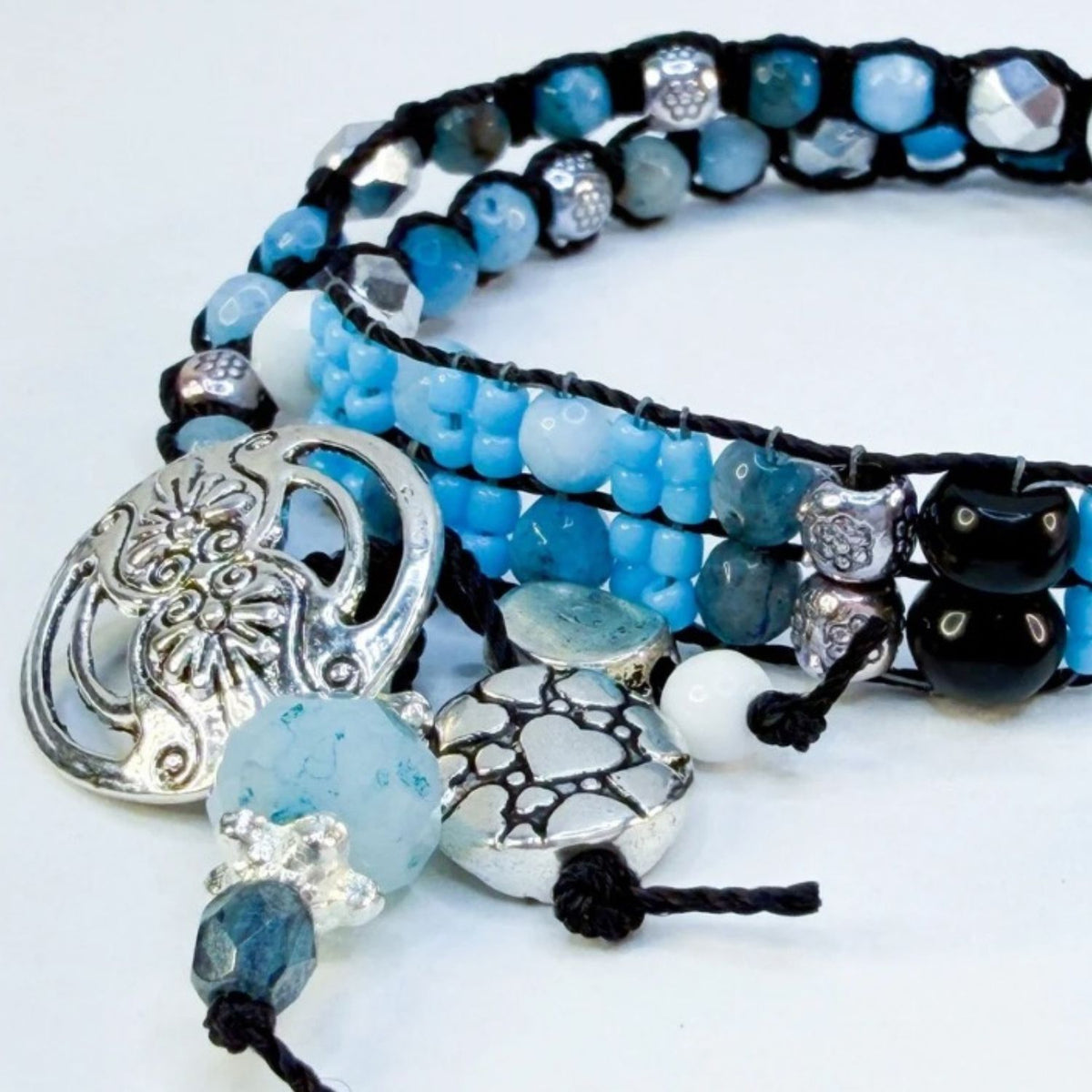 A close-up of a multi-strand beaded bracelet with light blue, dark blue, and white beads, black cord, silver-toned filigree charms, and faceted clear beads on a white background.