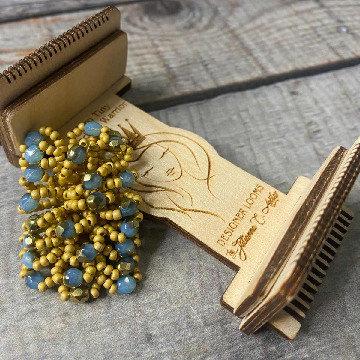 Earring Loom with example of what can be made featuring fire polish beads and seed beads in blue and yellow. Loom has the wisdom warrior artwork laser design on the front of the loom. The background is a faux wood in hues of blue grey.
